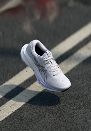 Weißer Laufschuh mit Schnürsenkeln, der über einer Asphaltstraße mit weißen Fahrbahnmarkierungen schwebt und darunter einen Schatten wirft.