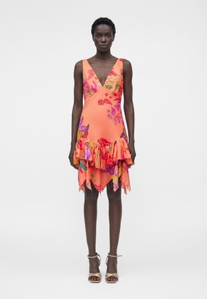 Woman wearing a sleeveless, orange floral dress with a deep V-neck and ruffled asymmetrical hem, paired with beige ankle-strap heels.