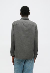 Chemise grise à manches longues avec un col, présentant une coupe droite, des poignets boutonnés et un tissu légèrement texturé. Portée avec un jeans bleu clair.