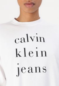 Balta medvilninė marškinė su juodu užrašu "calvin klein jeans". Turi apvalų kaklą ir laisvą kirpimą.
