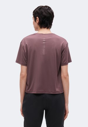 Personne aux cheveux courts et foncés portant un t-shirt ample mauve à manches courtes avec un texte vertical de logo "KARITRAA" dans le dos et un pantalon noir.