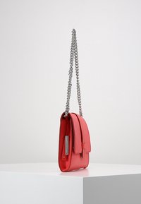 Sac à main en cuir rouge avec une bandoulière chaîne argentée, forme rectangulaire et rabat avant. Présente un détail logo métallique sur le côté.
