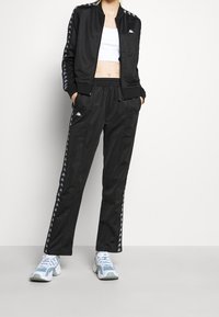 Svart träningsdräkt med zip-jacka och byxor med elastisk midja. Vit logotypdetalj på bröstet och randiga detaljer längs ärmarna och byxorna.