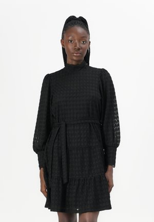 Black dress made of textured fabric, featuring a high neck, long puffed sleeves, a tiered design, and a waist tie for an adjustable fit.