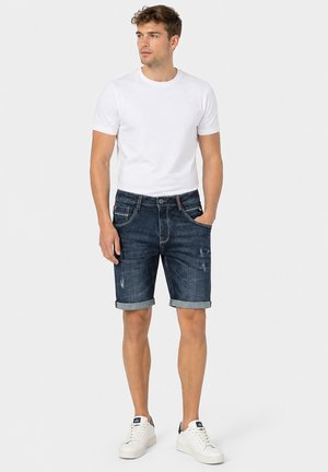Junger Mann trägt weißes T-Shirt mit Rundhalsausschnitt, dunkel, hochgekrempelte Jeansshorts und weiße Sneakers, steht mit einer Hand in der Tasche.