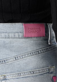 Lyseblå denimjeans med et rosa skinnmerke i livlinjen. Funksjoner inkluderer et tone-i-tone søm mønster og et lite brodert hjerte.
