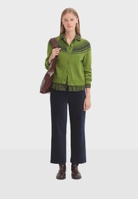 Groene gebreide cardigan met decoratieve blauwe accenten, knopen aan de voorkant en geribde manchetten, gecombineerd met een gestreept shirt en marineblauwe brede broek. Bruine tas en laarzen.