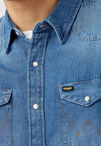 Jasnoniebieska koszula dżinsowa z kołnierzykiem w kształcie szpica, guziki na zatrzask, dwie kieszenie na piersi z wzorami stębnowania oraz metką z logo Wrangler.