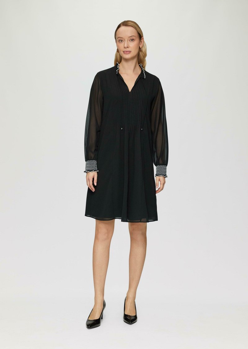 Schwarzes Kleid mit langen durchsichtigen Ärmeln, Steppdetails und gemusterten Manschetten. Der Kragen hat eine kontrastierende Einfassung. Knielanges Design.