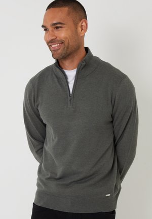 Mand smilende iført en mørkegrå sweater med kvart lynlås over en hvid skjorte, stående mod en ensartet lys baggrund.