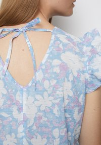 Hellblaues Blumen Kleid mit Puffärmeln, das einen tiefen V-Ausschnitt und einen Bindeverschluss hat. Der Stoff hat eine strukturierte Optik mit rosa Akzenten.