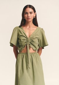 Olive zelena kratka majica s V-izrezom, kratkimi valovitimi rokavi, naborki spredaj in detajlom z zavezovanjem v pasu, združena z usklajeno krilo.