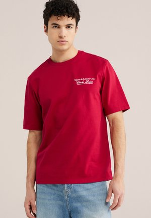 Mladić tamne kovrčave kose, nosi crvenu majicu kratkih rukava s bijelim tekstom na prsima i svijetloplave traperice na jednobojnoj pozadini.