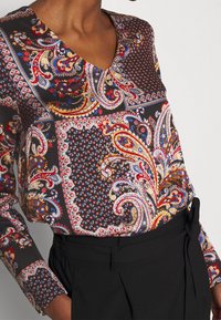 Langarmbluse aus gemustertem Stoff mit einem V-Ausschnitt. Die Farben umfassen Schwarz, Rot, Blau und goldene, filigrane Paisleymuster.