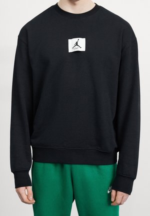 Person iført sort sweatshirt med lille hvid Jumpman-logo på brystet og grønne Nike joggingbukser.