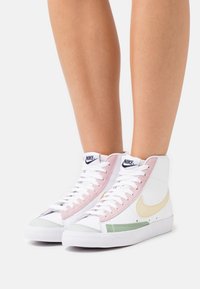 Wysokie trampki z białą skórzaną cholewką, różowymi i zielonymi zamszowymi akcentami oraz beżowym logo Nike. Posiadają białe sznurowadła i fakturowane podeszwy.