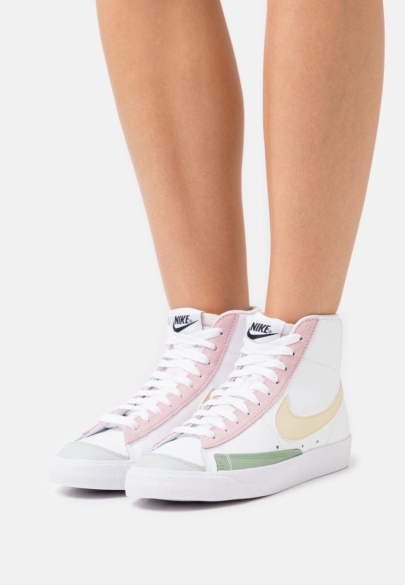 Wysokie trampki z białą skórzaną cholewką, różowymi i zielonymi zamszowymi akcentami oraz beżowym logo Nike. Posiadają białe sznurowadła i fakturowane podeszwy.