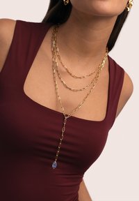Collar de cadena de oro con diseño en capas, que presenta eslabones alargados y un colgante con piedra preciosa azul. Llevado sobre una blusa burdeos.