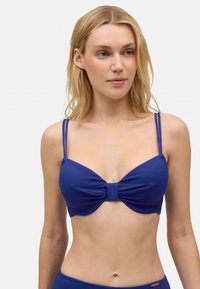 Top de bikini azul con tejido texturizado, detalle de lazo en el centro y finas tiras ajustables. Diseño llamativo con suaves curvas.