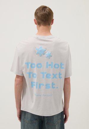 Joven visto de espaldas con una camiseta gris claro con texto azul "Too Hot To Text First" y dos formas de estrella sobre el texto.