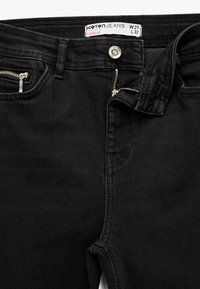 Los jeans de algodón negros tienen un bolsillo delantero estándar con cierre de cremallera plateada y botón. La etiqueta indica la talla W27 L32 y el estilo "Push Up".