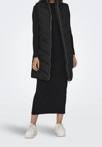 Svart pufferväst med hög krage och dragkedja, med en quiltad design och sidofickor, bärs över en svart lång klänning och vita sneakers.