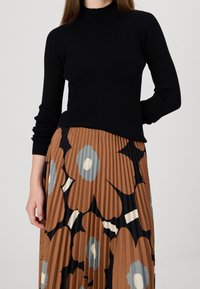 Donna che indossa un maglione nero a coste e una gonna marrone plissettata con grandi fantasie floreali blu e crema, in piedi con una mano dietro la schiena.