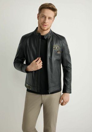 Mężczyzna w czarnej skórzanej kurtce z naszywkami, czarnej koszuli i beżowych spodniach, stojący na tle jasnoszarego tła.