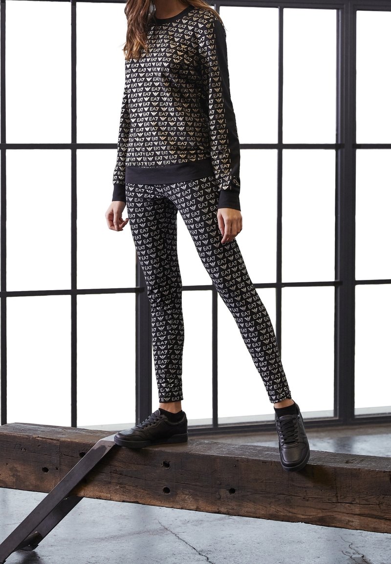 Černá mikina a legíny s opakujícím se vzorem loga značky ve zlaté barvě. V kombinaci s černými teniskami a umístěné před velkými okny.