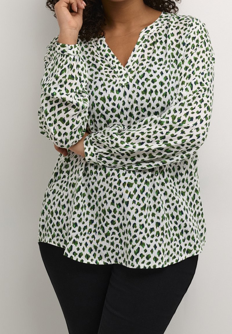 Femme portant une blouse blanche à manches longues avec des taches abstraites vertes et noires, associée à un pantalon noir, debout devant un fond uni.