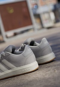 Šedé sportovní boty s texturovaným koženým svrškem, třemi bílými pruhy a gumovou podešví. Značka viditelná na patě a boku.