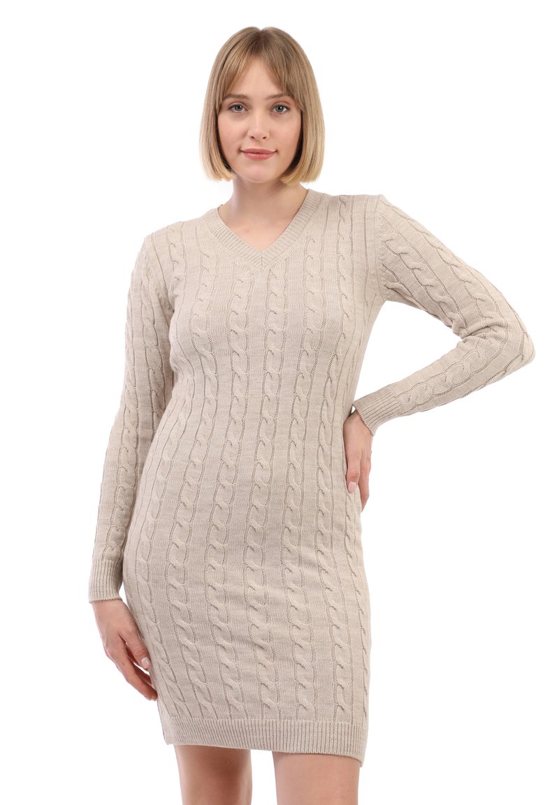Giovane donna con i capelli biondi corti che indossa un vestito maglione beige a trama intrecciata con scollo a V, posando con una mano sulla vita contro uno sfondo bianco.