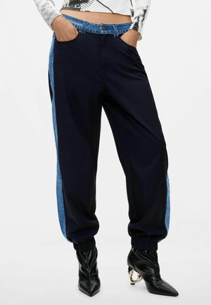 Donna che indossa pantaloni bicolore con davanti scuro, lati in denim, caviglie elasticizzate e stivali neri in pelle con tacco, in piedi con le mani nelle tasche.