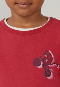 Czerwona bluza z białym ściągaczem przy dekolcie. Zawiera grafikę tygrysa jadącego na motocyklu, z napisem "BIG SHOW" w kolorze białym.