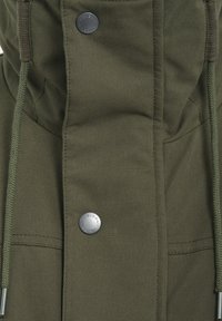 Chaqueta de tela verde olivo con cierre frontal que presenta botones plateados. Incluye drawstrings en la capucha y un material texturizado.
