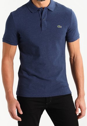 Man draagt een nauwsluitend marineblauw poloshirt met korte mouwen en een klein groen krokodillenlogo op de linkerborst.