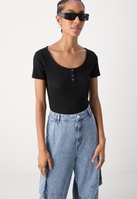 Cămașă neagră din bumbac, cu decolteu rotund și trei nasturi, combinată cu pantaloni cargo din denim texturat de culoare albastru deschis.