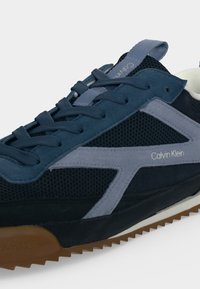 Baskets Calvin Klein bleu marine et grises avec dessus en mesh, surpiqûres en daim, lacets et semelle en caoutchouc marron à crampons.