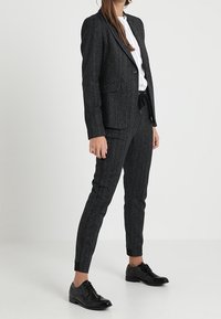 Černý pruhovaný oblek se střiženým sakem a slim-fit kalhotami, vyrobený z texturované látky. Nošený s bílou košilí a černými botami.