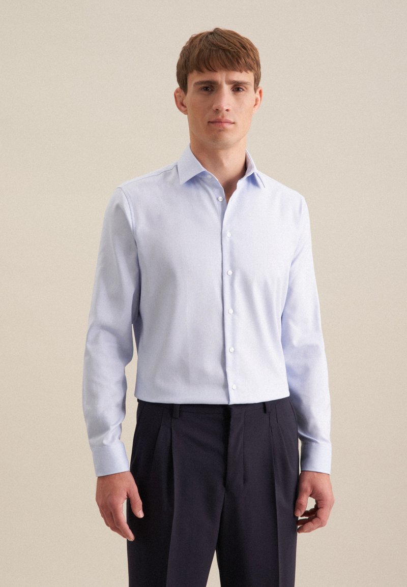 Jeune homme portant une chemise habillée bleu clair boutonnée et un pantalon sombre à plis, debout devant un fond beige uni.