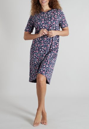 Woman wearing a navy blue knee-length dress with short sleeves and a pink and white floral pattern, standing barefoot on a plain background.