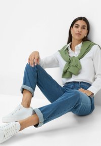 Weißes Hemd mit Knopfverschluss, grüner Pullover um den Hals gebunden, blaue Jeans mit hochgerollten Säumen, weiße Sneaker, glattes Material und lässige Passform.