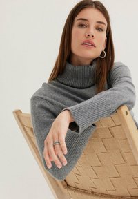 Pull en col roulé gris à côtes avec une coupe ample, associé à une chaise en osier. Comprend des manches longues et une texture visible. Bague en argent simple au poignet.