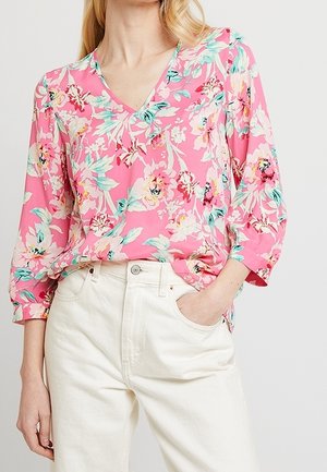 Kvinde iført en lyserød bluse med lange ærmer og pastelblomstret print, stoppet ned i højtaljede, offwhite bukser, med den ene hånd i lommen.