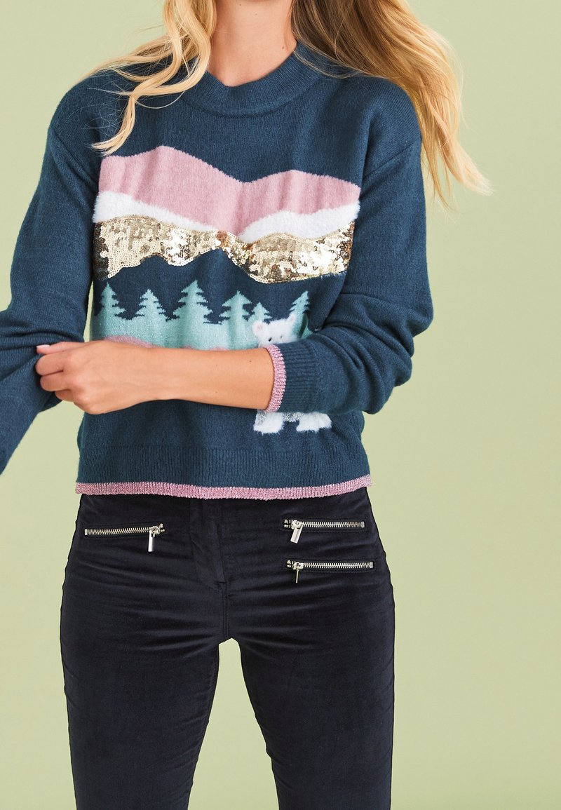 Femme portant un pull bleu marine avec des motifs de montagnes et de forêt en rose, blanc et or, incluant un petit ours, associé à un pantalon noir avec quatre fermetures éclair.