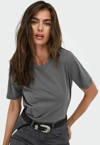Camiseta gris de manga corta en tejido suave, con cuello redondo, combinada con un cinturón negro con una hebilla decorativa de plata, llevada con pantalones vaqueros oscuros.