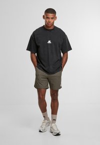 Mladý muž stojí s rukama v kapsách, oblečený v černém oversized tričku, olivových kraťasech, béžových ponožkách a stříbrných běžeckých botách na bílém pozadí.