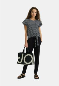 T-shirt a righe bianco e nero con nodo frontale abbinato a pantaloni neri e una grande borsa tote con grafica "JO". Sandali neri completano l'outfit.