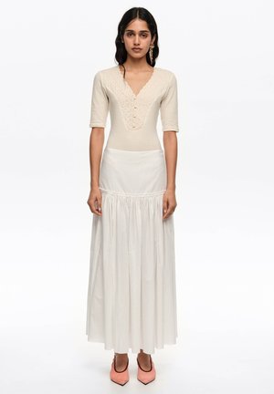 Mujer con una blusa beige de manga corta con detalles de encaje, una falda larga blanca de cintura alta y tacones rosas de punta puntiaguda, posando frente a un fondo blanco.