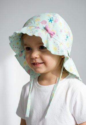 Toddler smiling, wearing a white shirt and a pastel sun hat with sea-themed patterns and a pink bow, against a plain light background.